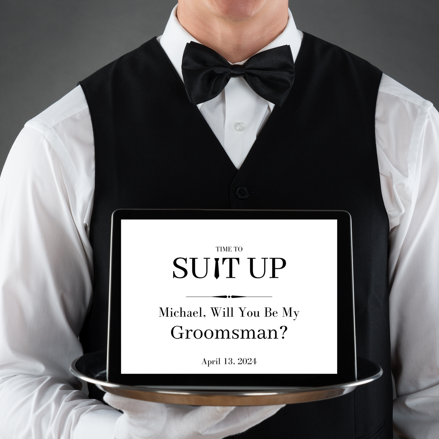 Man in tuxedo holding a tray with a 'Will You Be My Groomsman?' proposal on a screen.