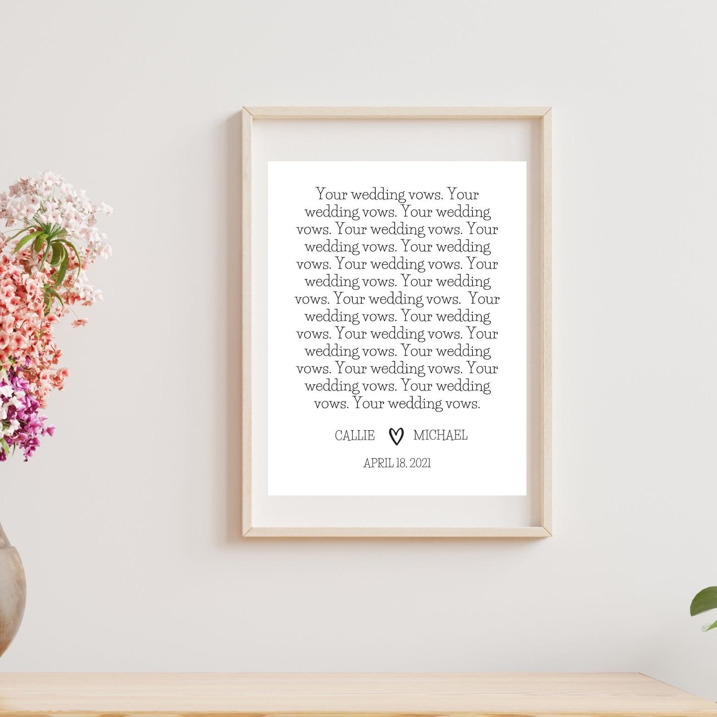 A customizable wedding vows print featuring text in a decorative font, displayed in a frame on a wooden surface, with a potted plant and a vase with flowers to the left.