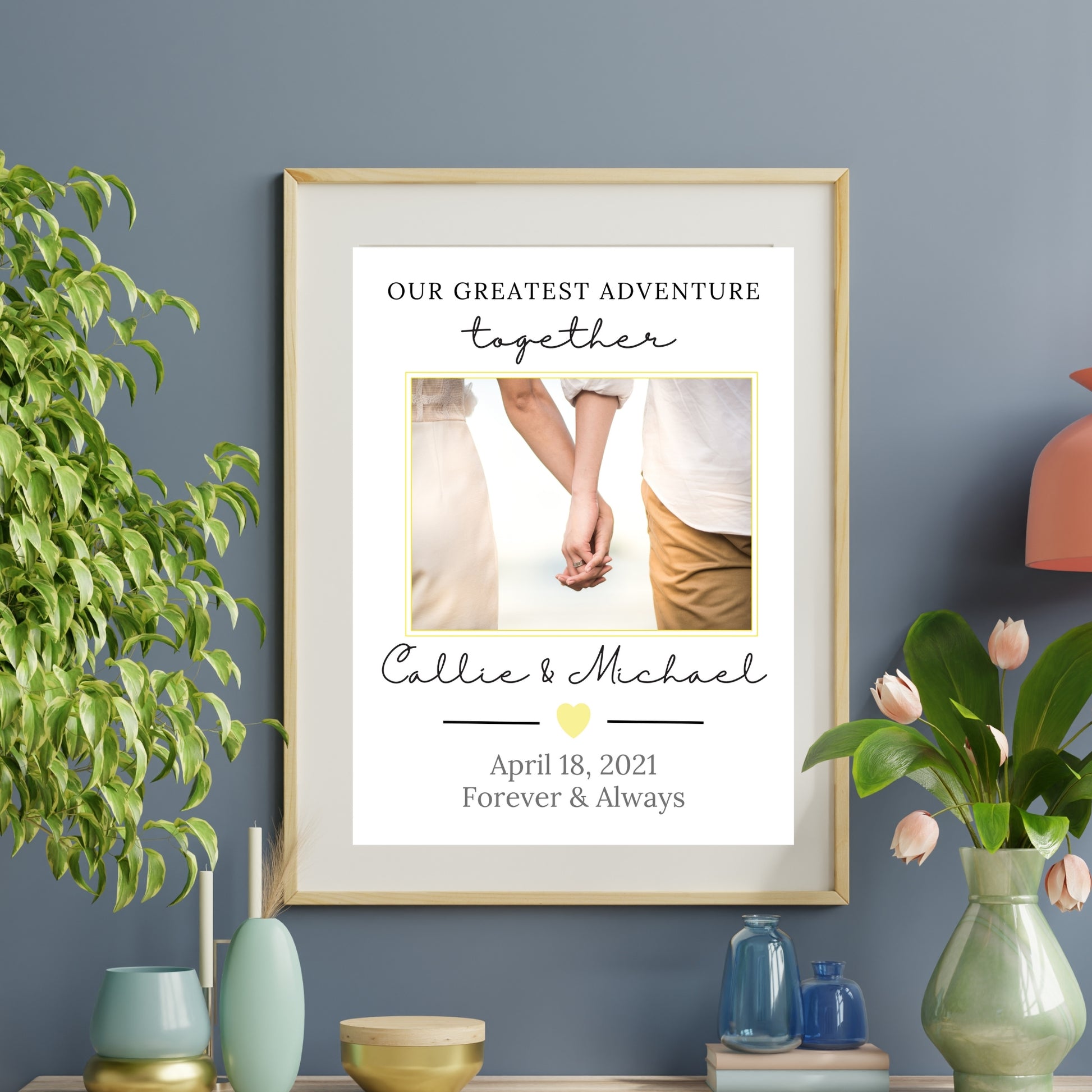 Framed print with 'Our Greatest Adventure Together' text and a couple holding hands, on a wall with decorative vases and plants.
