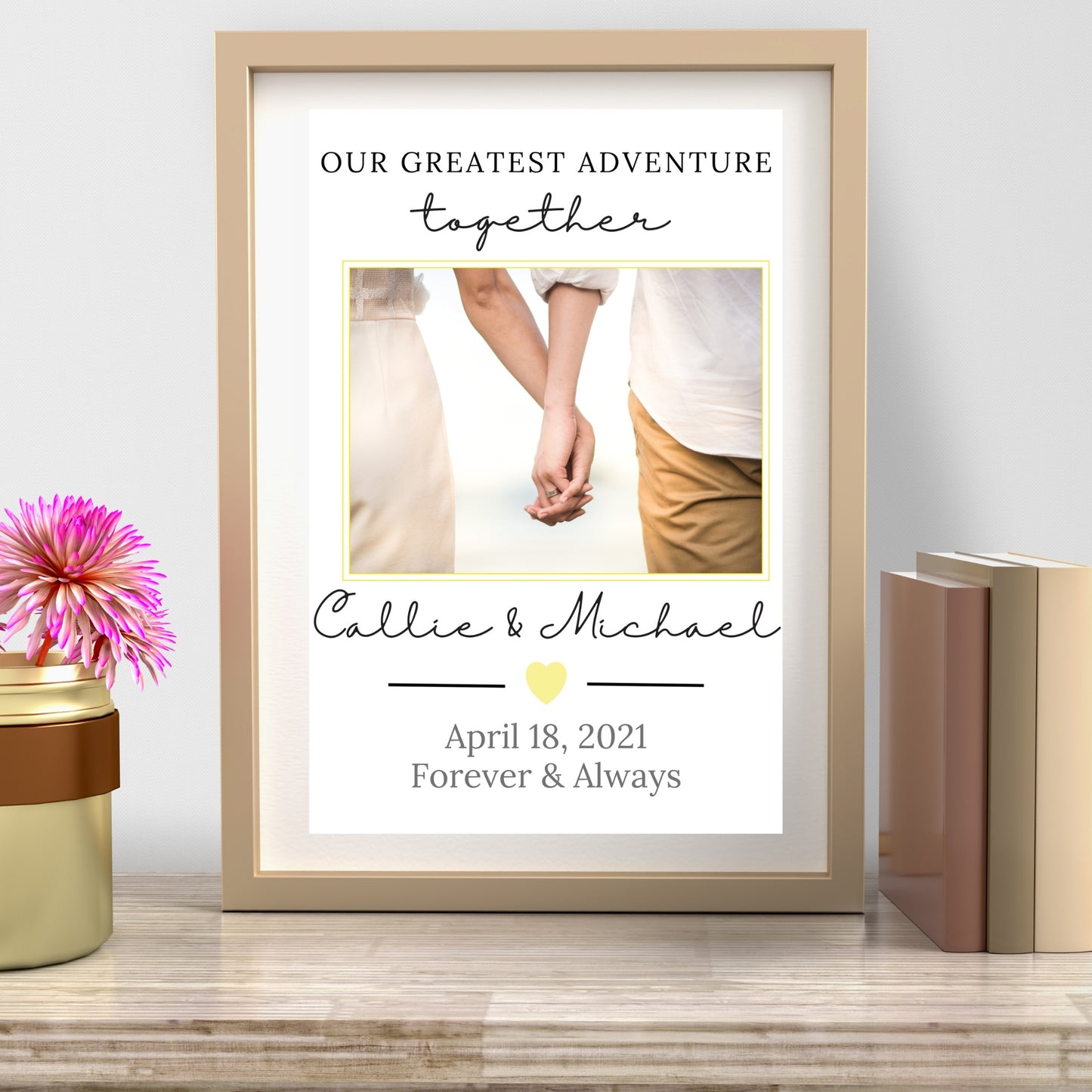 Framed print with a couple holding hands, personalized text, and a pink flower on a wooden surface.