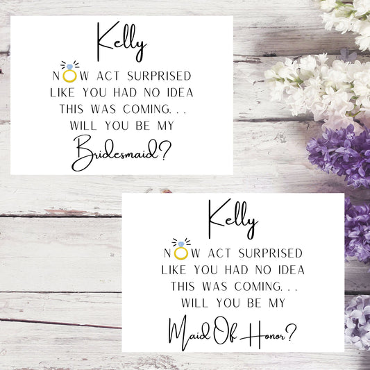 Two cards with personalized requests for bridesmaid and maid of honor on a wooden surface with flowers.