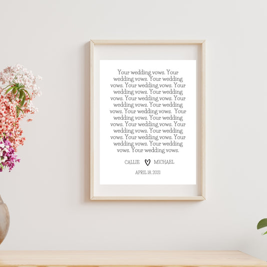 A customizable wedding vows print featuring text in a decorative font, displayed in a frame on a wooden surface, with a potted plant and a vase with flowers to the left.